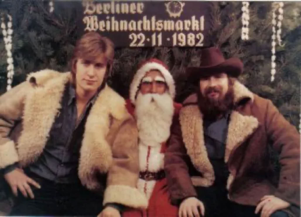 Drei Personen sitzen vor einem Schild mit der Aufschrift "Berliner Weihnachtsmarkt 22-11-1982", in der Mitte eine Person in einem Weihnachtsmannkostüm, die anderen beiden tragen Pelzmäntel.
