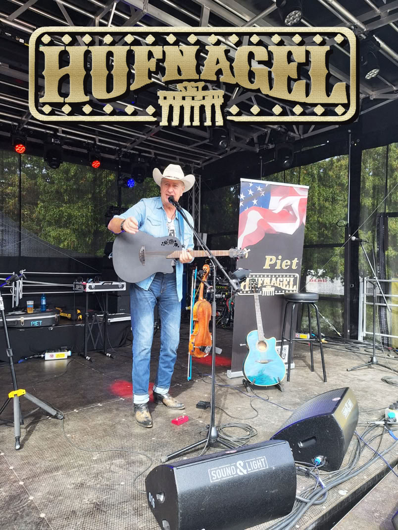 Ein Musiker mit Cowboyhut spielt Gitarre auf einer Bühne. Im Hintergrund sind ein Banner mit der Aufschrift "Hofnagel" und ein Plakat mit einer amerikanischen Flagge zu sehen. Auf der Bühne stehen weitere Instrumente und Ausrüstung.