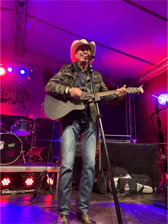 Ein Musiker mit Cowboyhut spielt Gitarre auf einer Bühne mit bunten Lichtern im Hintergrund.