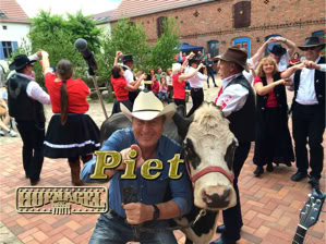 Eine Gruppe von Menschen in traditioneller Kleidung tanzt im Freien, ein Mann mit Cowboyhut posiert mit einer Kuh im Vordergrund.