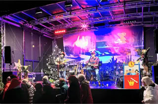 Eine Bühne mit Musikern, die Instrumente spielen, umgeben von Weihnachtsdekorationen und einem Publikum im Vordergrund.