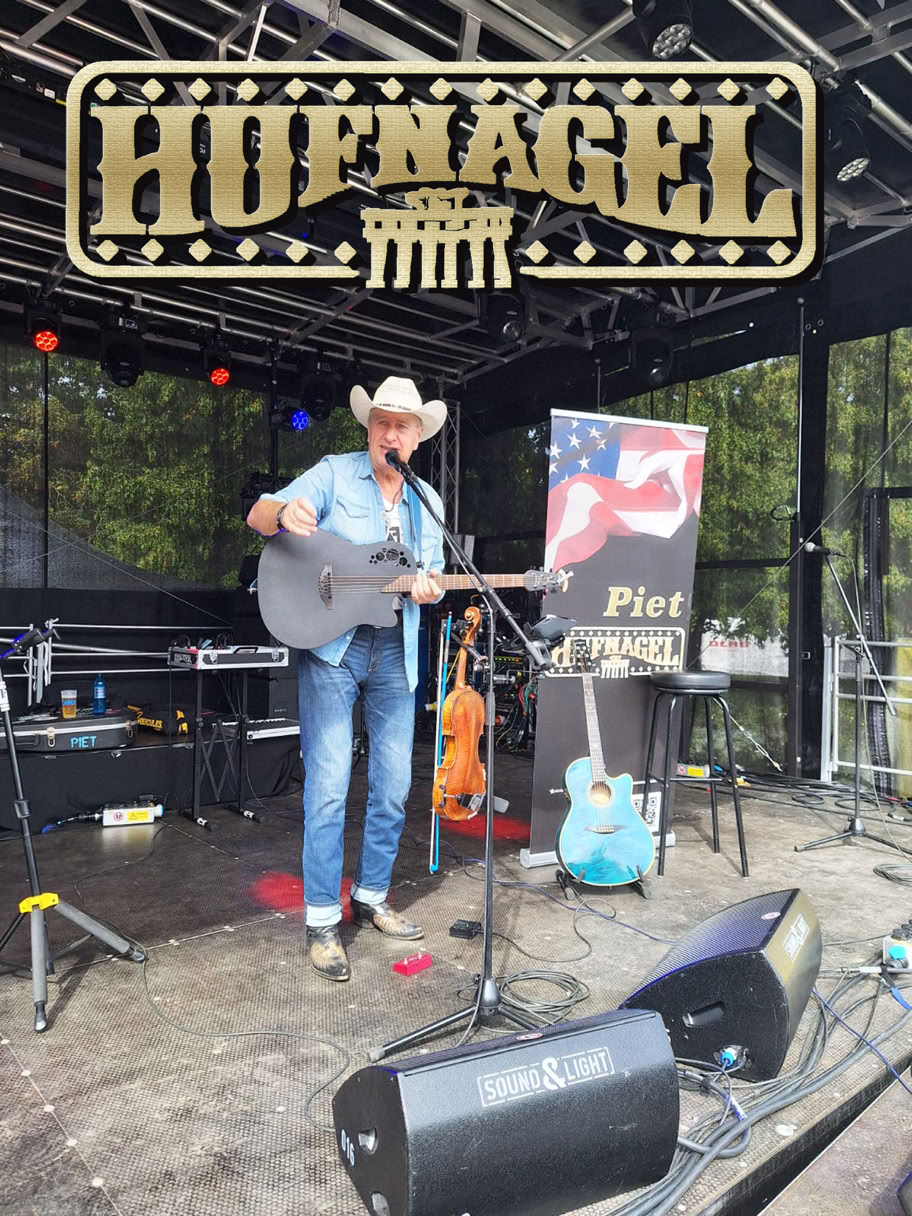 Ein Musiker mit Cowboyhut spielt Gitarre auf einer Bühne. Im Hintergrund sind ein Banner mit der Aufschrift "Hofnagel" und ein Plakat mit einer amerikanischen Flagge zu sehen. Auf der Bühne stehen weitere Instrumente und Ausrüstung.