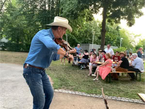 Ein Mann mit Cowboyhut spielt Geige im Freien, während eine Gruppe von Menschen an Tischen im Hintergrund sitzt.